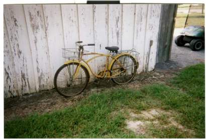 ellow bike with front and back baskets leans against a worn white wall, in the grass.